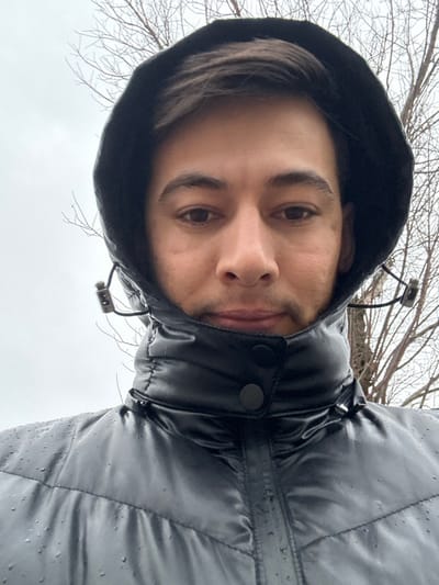 A male individual is depicted in a close-up, head-and-shoulders portrait, consistent with a selfie, located in Yangiiul, Uzbekistan. He has short, dark hair visible under a pulled-up, black hooded puffer jacket. His gaze is directed forward. The jacket features a high collar secured by two black snap buttons and shows numerous water droplets on its shiny, possibly water-resistant surface, particularly across the chest and shoulders. Drawstring adjusters with metallic stoppers are visible on the sides of the hood. The background consists of a uniformly overcast, light grey sky and a scattering of leafless tree branches, suggesting a cold and wet environment.