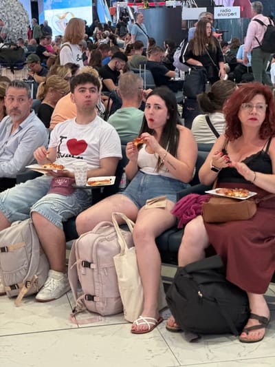 The image depicts a crowded waiting area, likely in an airport, with numerous people seated or standing. The setting suggests the city of Fiumicino, Italy, given the presence of individuals eating pizza and wearing "Italia" branded clothing.

Several individuals are prominently featured in the foreground. A woman with red hair is seated and appears to be holding a slice of pizza. Next to her, a woman with dark hair is also eating pizza. A young man in a white t-shirt with a red heart graphic sits alongside them, also eating pizza. Further down in the image, an older man in a light blue shirt is also eating a slice.

The environment is filled with people, and the lighting suggests an indoor location. Luggage, bags, and personal belongings are scattered around the seating area.