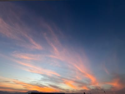 This media file captures a beautiful and expansive sky scene, most likely at sunset or sunrise, over San Francisco Ocotlán, Mexico. The dominant subject is the sky, featuring a captivating gradient of colors. The upper portion of the sky is a deep, rich indigo blue, gradually fading into lighter shades of blue and purple towards the middle.

Below this, wispy, elongated clouds are illuminated with warm, vibrant hues. These clouds are predominantly pink, orange, and peach, appearing in streaky formations that stretch across the frame, especially prominent on the left side where they seem to radiate upwards. The intensity of these warm colors is strongest in the lower half of the sky, blending into lighter, softer tones.

At the very bottom of the image, a dark, silhouetted horizon line is visible. On the left, this silhouette suggests distant mountains or hills. On the right, faint dark shapes indicate structures, possibly rooftops or buildings with antennas and poles, against the glowing sky. A very thin, dark line, possibly a wire or edge of an overhead structure, runs horizontally along the very top edge of the frame.

The overall scene conveys a sense of tranquility and natural beauty, characteristic of dawn or dusk. There are no visible people or discernible text. The weather appears clear with high-altitude cloud formations, contributing to the dramatic sky display.