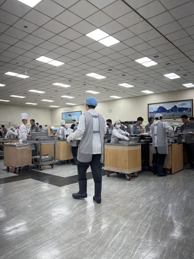 The image depicts an indoor dining or food service area, likely a cafeteria or mess hall, within a facility in Geoje-si, South Korea. The space is illuminated by an overhead grid of fluorescent ceiling lights. Several individuals, primarily wearing white chef hats or light-colored caps and aprons, are engaged in food service activities. Some are preparing or serving food from large metal trays and containers mounted on wheeled carts. The floor is tiled with a gray marble-like pattern, with darker inlaid stripes. In the background, a framed picture of a mountainous landscape with floral elements hangs on the wall. The overall atmosphere suggests a functional, organized environment for mass food distribution.