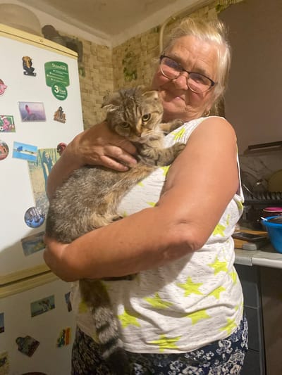 A woman with blonde hair and glasses is holding a tabby cat. She is wearing a white t-shirt with yellow stars and dark patterned pants. The background shows a kitchen with a refrigerator covered in magnets and a counter with dishes. The image appears to be taken indoors, possibly in a residential setting. The city of Маринопіль, Ukraine is mentioned, but there is no visual information in the image to confirm or deny this location.