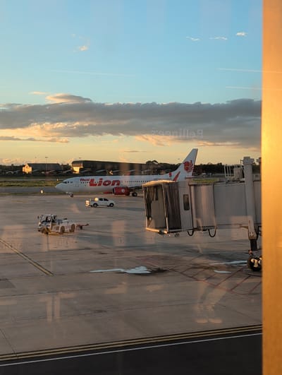 An airplane with "Thai Lion Air" branding is parked on the tarmac at the airport. A jet bridge is connected to the aircraft. A small white vehicle is positioned in front of the plane, and a tow tractor is visible nearby. The scene appears to be in Tambon Hua Toei, Thailand. The sky above is a clear blue with scattered clouds, and the lighting suggests it is either sunrise or sunset. The ground is paved with asphalt and marked with yellow and white lines. There are reflections on the paved surface, possibly from water or oil. In the background, several airport buildings and structures are visible.