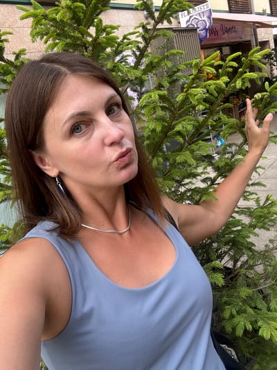 A woman with brown hair, wearing a light blue tank top and a silver necklace, poses for a selfie outdoors. She is positioned in front of a large green coniferous tree, with its branches extending across the frame. In the background, a building with signage for "FLORISTE" and "Miss Vanda Artisan Fleuriste" is visible. The woman is puckering her lips and her right arm is bent, with her hand near the tree's branches. The overall scene suggests an urban environment, possibly a street in Les Rousses, France, given the florist shop.