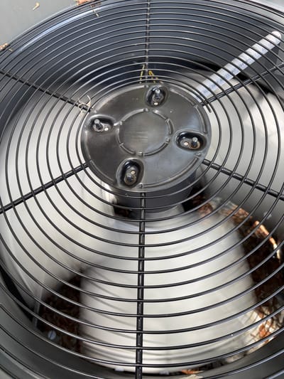 The image shows a close-up, slightly angled view of the fan grill of an outdoor air conditioning unit. The fan blades themselves are blurred due to motion, indicating the unit is operating. The grill is made of black metal wires forming concentric circles and radial supports. In the center of the grill is a black, circular housing with three visible bolts and a smaller, off-center circular element with two visible bolts, likely covering the motor.
Scattered debris, appearing as small brown organic matter like leaves or twigs, is visible on the grill and slightly behind it, caught within the fan assembly. A thin, white, cylindrical object with black text is partially visible through the grill on the left side of the image. The text is difficult to read entirely, but "SULIGT R R515RT CNPRHS TD E G3 CS" is discernible.
The scene is lit by natural light, suggesting it is daytime. The setting appears to be outdoors, consistent with an air conditioning unit. The overall impression is one of a functional, outdoor appliance in operation, with some minor signs of environmental exposure. No people are visible. The location is specified as Bradenton, United States.