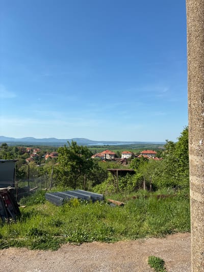 The image shows a view in Bokilovtsi, Bulgaria. The sky is clear and blue. The foreground features a concrete structure and overgrown grass. In the distance, there are houses with red roofs amidst green vegetation, suggesting a village setting. Hills or mountains are visible in the far background. Wire mesh and wooden planks lay in the grass.