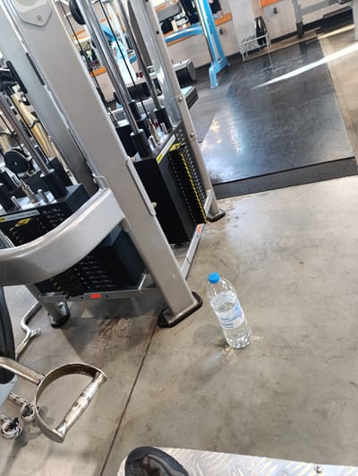 The image presents an indoor scene within a gym or fitness center located in Rakovski, Bulgaria. The primary focus is on the lower section of a large cable weight machine, characterized by its sturdy silver-gray metal frame and multiple black weight stacks.
In the immediate foreground, a person's foot, clad in a dark athletic shoe, is partially visible on a silver, diamond-plate textured platform. On the concrete floor, next to the weight machine, lies a chrome D-handle exercise attachment and several coiled rope attachments. A clear plastic water bottle with a blue cap and a label, part of which reads "Buaro" (likely a brand name like "Aquaro"), stands upright on the concrete. The floor shows some marks and general wear, typical of a busy gym environment.
The visible weight stack displays various weight increments in both pounds and kilograms, such as "22 lb / 10 kg," "33 lb / 15 kg," "44 lb / 20 kg," and continues with higher values like "187 lb / 85 kg," "209 lb / 95 kg," "231 lb / 105 kg," "253 lb / 115 kg," "275 lb / 125 kg," and "297 lb / 135 kg." A warning label on the equipment states "TION Do Not Alter Or Modify Weight Stack."
In the background, beyond the machine, a dark, rubberized floor mat transitions into another concrete area where other gym equipment, including part of a blue-framed apparatus and a rack with weight plates, can be discerned. A bright stripe of light, possibly from a window or overhead lighting, crosses the floor in the mid-background. No people are visible, but the setup, equipment, and water bottle suggest that a workout session is either underway or has recently concluded. The lighting indicates an indoor setting, likely during daylight hours.