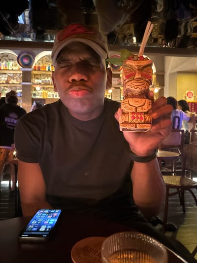 An indoor, dimly lit scene captures a person holding an elaborate tiki cocktail mug, likely at a bar or restaurant in Longta, China. The image is oriented vertically but appears rotated 90 degrees to the left.
In the foreground, a dark-skinned individual with a slight mustache, wearing a dark t-shirt and a light-colored headband over a reddish-pink band, looks directly at the camera with a neutral expression. They are holding up a decorative tiki mug with both hands. The mug is intricately carved with facial features and geometric patterns in beige and brown, accented with red, and features two straws and a green leaf garnish.
To the right of the person, on a dark table, lies a smartphone with its screen illuminated, displaying app icons and the time "09:07". A round coaster, partially visible, has text that reads "HORN..." and appears to be a brand related to "TEQUILA."
The background is softly blurred, revealing other patrons and the interior decor of what appears to be a themed establishment, possibly a tiki bar. Shelves filled with bottles and various decorative items, including a colorful circular design, are visible. The ambient lighting is warm, suggesting an evening setting. Another person in the background wears a dark t-shirt with a partial logo "HORNW...". A red sign with "30" in a circle is also faintly visible in the far background. The activity suggests a social outing, with the main subject posing with their drink.