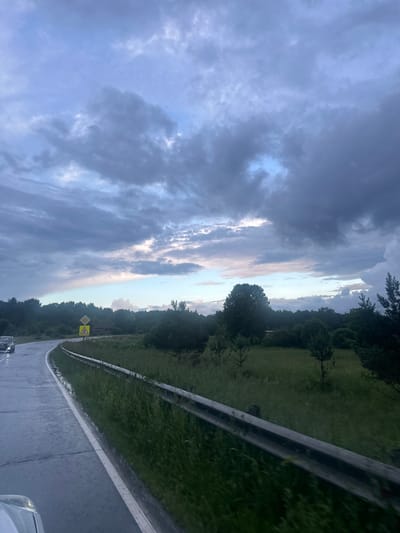 This is a vertical, slightly tilted, eye-level shot of a road with a guardrail and a grassy embankment under a dramatic, cloudy sky. The sky is filled with dark, heavy clouds, with patches of lighter sky showing through, suggesting an approaching or receding storm. The time of day appears to be either early morning or late evening due to the dim lighting and the colors in the sky.
The scene is a rural roadside, with lush green grass and trees lining the embankment. A road, appearing wet as if from recent rain, curves to the right and is bordered by a metal guardrail. There are two yellow diamond-shaped warning signs visible on the side of the road. The first sign has a symbol of a pedestrian crossing, and the second sign has a symbol of a warning triangle, indicating a potential hazard ahead.
The overall mood is somewhat somber and atmospheric, due to the dark clouds and the wet road. There are no people or animals visible in the frame, but the presence of the road and guardrail suggests it is a passage for vehicles. The location context is Uva, Russia, a setting that can be associated with this type of landscape.