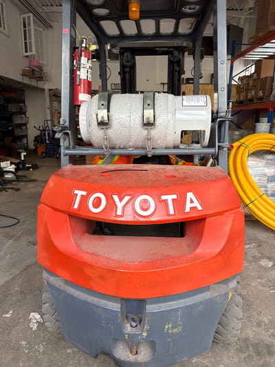 This is a full-frame, eye-level, forensic, matter-of-fact photograph of the front of an orange and grey Toyota forklift. The forklift is parked indoors on a concrete floor. In the foreground, the word "TOYOTA" is prominently displayed in white lettering on the orange body of the forklift. A grey, ribbed panel is visible below the lettering. The forklift has large, grey tires.

Mounted on the mast of the forklift is a white propane tank wrapped in bubble wrap, secured by metal brackets. A red fire extinguisher is attached to the mast to the left of the propane tank. A yellow coiled hose is visible on the right side of the image, in front of a metal shelving unit stacked with boxes.

In the background, various items are visible, including another forklift, shelves filled with supplies, and a window. The overall environment suggests a warehouse or industrial setting. The image does not provide specific information about the city of Richmond Hill, Canada.