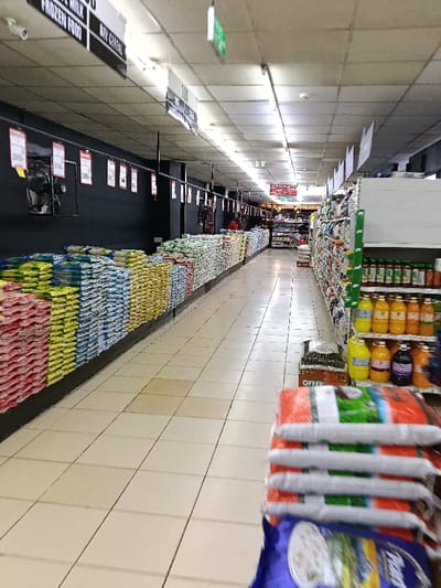 The image captures a long, well-lit aisle within a supermarket or large grocery store. The perspective is looking straight down the aisle, with items stacked high on either side.
On the left side of the aisle, a long display runs the entire visible length, consisting of numerous large bags of goods stacked neatly in colorful rows. These bags appear to contain bulk items such as rice, flour, or other dry provisions. The colors of the bags alternate, showing sections of red, yellow, blue, and white/green packaging. Behind these stacked bags, the wall is painted a dark color, possibly black or dark grey. Attached to this wall are several red price tags with white numbers, and some framed posters or advertisements. Visible price tags include "295/-", "319/-", and "269/-" further down the aisle. Hanging from the ceiling are overhead signs indicating product categories. From left to right, these signs read "FROZEN FOOD", "MILK", and "DRY CEREAL". A small white security camera is also visible on the ceiling near the "FROZEN FOOD" sign.
On the right side of the aisle, there are typical supermarket shelves displaying a variety of products. The lower shelves are clearly stocked with numerous bottles of liquids, appearing to be juices or other beverages, in various colors such as yellow, orange, and dark purple/blue. Further up the shelves, other packaged goods are visible, though less distinctly.
The ceiling consists of light-colored, possibly acoustic, tiles with long fluorescent light fixtures providing bright illumination to the aisle. The floor is covered with large, light-colored (beige or cream) square tiles with dark grout lines, showing some signs of wear or minor discoloration in places.
Towards the far end of the aisle, the store continues, with more shelves and displays visible. A person wearing a red top is faintly visible in the distance, near the end of the left-side display. A red sign is hanging further down the aisle, but the text is blurry; it might be "CUSTOMER SERVICE" or a similar store section indicator.
In the immediate foreground, at the bottom right of the image, several more large bags of product are visible, stacked on top of each other. These bags are somewhat blurry, suggesting they are very close to the camera or in motion, possibly indicating the viewpoint from a shopping cart. One blue bag at the very bottom has partial text visible, which appears to include "PLANT". Next to these bags, a cardboard box is visible with the word "OFFER" printed on it.