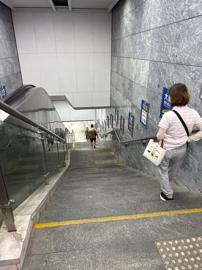 A downward-facing, wide-angle shot captures a scene within a modern subway station in Liwan District, China. A woman, viewed from behind, descends a wide, grey stone staircase. She wears a light pink striped polo shirt, grey sweatpants, and white gloves, carrying a small, cream-colored tote bag adorned with cartoon bears. Her black and white sneakers are visible as she walks. To her left, a glass-paneled escalator railing curves upwards. Further down the stairs, two men walk in the same direction. The station walls are clad in grey, horizontal stone slabs. Blue signage with white Chinese characters is mounted on the wall to the woman's right. A bright yellow line is painted on the edge of the stairs, marking a safety boundary. The lighting is bright, typical of an indoor public space.