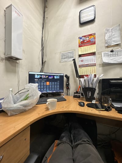 The image depicts an office workspace with a desk, computer, and various office supplies. The desk is made of wood and has a curved shape. On the desk, there is a computer monitor displaying a menu with icons, a keyboard, a mouse, a white mug with a tea bag string hanging out, a plastic bag containing some items, a pen holder filled with papers, a printer, a glass mug with liquid, and a smartphone. In the background, there is a wall with several calendars and papers attached. A white rectangular device is mounted on the wall to the left. The scene appears to be an indoor office environment. The image does not provide any specific details about the city of Sakmary, Russia, or its location within it.