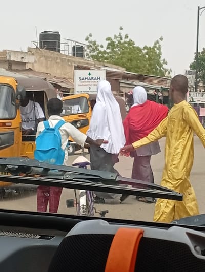 The image depicts a street scene in Hotoro North, Nigeria, viewed from inside a vehicle with the dashboard and windshield wipers visible in the foreground. The mid-ground features five individuals and multiple vehicles.
On the left, a person wearing a white t-shirt with "BOSS" printed on it is partially visible inside a yellow tricycle. Immediately to their right, a male, appearing to be a youth, wears a light-colored long-sleeved shirt, maroon-colored pants, and a bright blue backpack with a light blue circular emblem. He walks towards the right, extending his right hand to hold a white, conical object.
Further right and behind this individual, two females walk away from the camera towards the right. The first wears a white head covering and a dark, ankle-length garment. The second wears a white head covering, a prominent red shawl over a grey top, and a dark skirt. On the far right, another male in a long, flowing yellow traditional garment also walks away from the camera, his right arm slightly extended.
Another yellow tricycle is visible in the mid-ground, behind the walking individuals. The background consists of various buildings, some with corrugated metal roofs. A multi-story building features two black water tanks on its rooftop, supported by metal framing, with green foliage visible behind it. A white sign for "SAHARAH MEDICINE STORE & DIAGNOSTIC CENTER, NO. 138 HOTORO TSANYA" is prominent on one of the buildings. The sky is overcast.