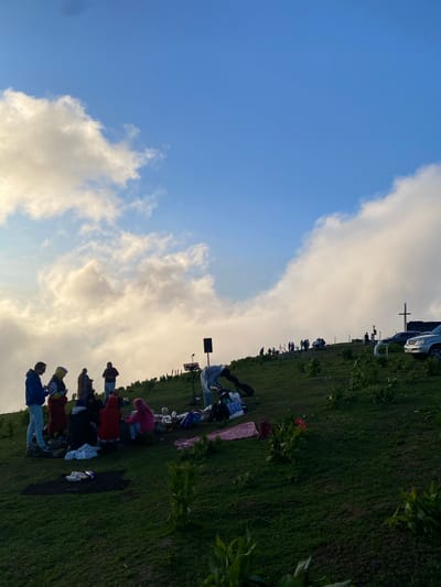 In Gomismta, Georgia, a group of people are gathered on a grassy hillside under a bright blue sky with scattered clouds. Some individuals are standing and talking, while others are seated on blankets, appearing to be having a picnic. There are several vehicles parked on the upper part of the hill, and a cross-shaped monument is visible in the distance. The landscape is characterized by rolling green hills and a few scattered plants.