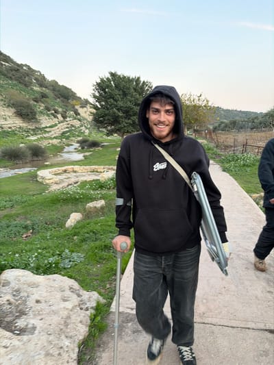 A young adult male, light-skinned with dark hair, walks on a paved pathway in the Emek Izrael Regional Council, Israel, smiling towards the camera. He wears a black hooded sweatshirt with a white logo, dark jeans, and dark sneakers. He uses a grey single crutch in his right hand for support. A grey, rectangular, folded object, possibly a portable chair, is slung over his left shoulder via a beige strap. Grey tape is visible on his left sweatshirt sleeve. To the left of the path is a grassy area with light-colored rocks, white flowers, and a winding stream. Beyond the stream, a steep hillside with exposed layered rock formations and sparse vegetation ascends. To the right of the path, a partially visible second individual's lower legs and a cultivated field with trees are present. The sky above is light blue.