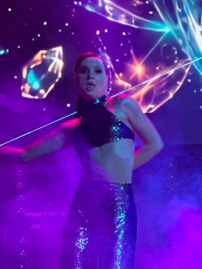 A woman in a sequined outfit performs on stage in Tsilkani, Georgia. The stage is lit with purple and blue lights, and a laser beam cuts across the scene. The backdrop is a projection of abstract, glowing shapes and stars. The woman is wearing a halter-neck crop top and a long sequined skirt. Her hair is pulled back, and she has bold makeup on. She is in motion, with her arms outstretched and her body turned. In the lower left corner of the image, the text "MOTIF X" is visible on a surface, likely a piece of equipment on stage. The overall atmosphere is vibrant and energetic, suggesting a musical performance or dance show.