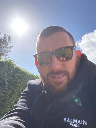 A man is taking a selfie outdoors in what appears to be a residential area, possibly Bondy, France. The sun is bright in the sky, casting a lens flare on the image. The man is wearing sunglasses and a black hoodie with "BALMAIN PARIS" logo on it. He has a beard and is smiling. In the background, there is a green hedge and a clear blue sky with some clouds. The man is holding his phone in his left hand to take the picture. The phone's reflection is visible in the man's sunglasses. The overall setting is a sunny day with good visibility.