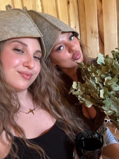 Two women are posing for a photograph in a sauna. Both women are wearing sauna hats. The woman in the foreground is fair-skinned with light brown hair and a gold necklace. The woman in the background is also fair-skinned with light brown hair and is holding a bundle of leafy branches and a black container. The sauna has wooden paneling. The location is unknown.