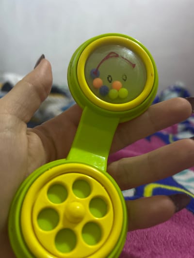 A hand with brown nail polish holds a green and yellow plastic toy with circular ends. One end has a clear cover with a smiley face and small, colorful balls inside. The other end has a yellow center with six holes. The background is blurry but includes a white surface and colorful fabrics. The location is unspecified and cannot be determined from the image.