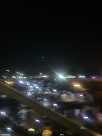 The image depicts a night scene in Sauka, Nigeria, characterized by blurred lights and indistinct forms, suggesting motion blur or a low-light setting. The foreground features a dark, sloping structure, possibly a railing or barrier. Beyond this, a multitude of colorful, elongated light streaks, some appearing as crescents, are scattered across the scene, indicating a busy urban environment with moving lights, such as vehicles or streetlights. Some areas show clusters of what appear to be stalls or displays with varying colors, hinting at a marketplace or commercial area at night. A faint, pale green arc is visible in the upper portion of the sky, which could be a sliver of the moon or atmospheric light. The overall impression is one of a vibrant, active night in the city, albeit rendered abstractly due to the photographic effects.