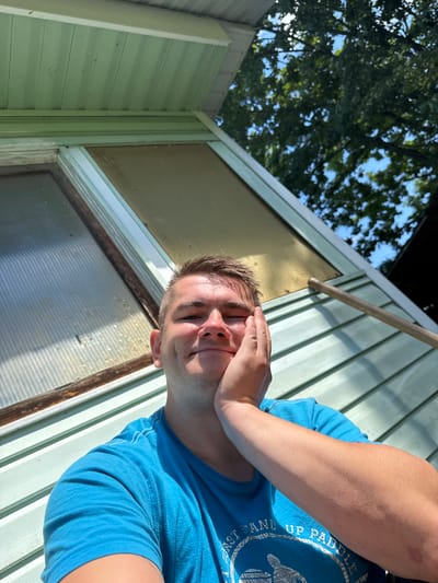 A young Caucasian male with short brown hair and fair skin is taking a selfie outdoors. He is wearing a bright blue t-shirt and has his right hand resting on his cheek, with his eyes closed and a slight smile. The background features a light green, clapboard-sided building with a window, a portion of a roof, and lush green trees against a blue sky. The scene appears to be in a residential area, likely a backyard, possibly located in the city of Krasnoe (Uleiminskoe S/P), Russia.