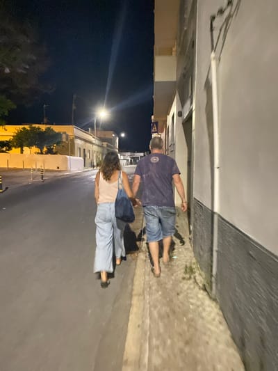 A couple, a man and a woman, walk hand-in-hand down a street in Olhão, Portugal at night. The man, wearing a dark t-shirt and denim shorts, walks barefoot, his t-shirt displaying a circular design on the back. The woman, in a light-colored tank top and wide-leg denim pants, also walks barefoot, carrying a dark blue tote bag. They are walking on a sidewalk next to a building with a grey, textured lower wall and a lighter upper section. Across the street, buildings with yellow facades and trees are visible under streetlights. The street appears wet, possibly from recent rain, and the sky is dark. The scene suggests a casual evening stroll through the town.