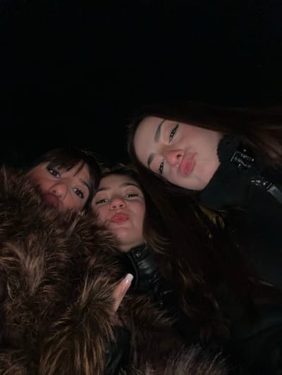Three young women pose closely together, angled downward toward the camera. The woman at the top has long, dark hair and wears a black jacket. The woman in the middle has similar hair and also wears a dark jacket. The woman at the bottom, closest to the viewer, has dark bangs and is surrounded by a large, brown fur collar or coat. All three women are making slightly puckered facial expressions, almost like they are blowing kisses. The background is black and out of focus.