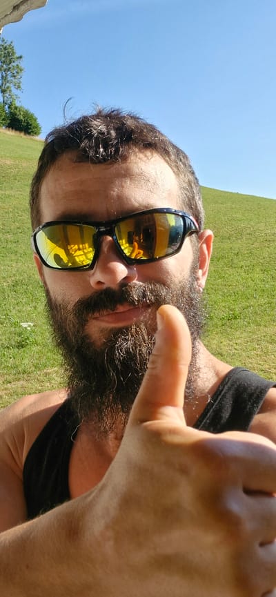 A man with a beard and sunglasses gives a thumbs-up while standing in front of a grassy hill on a sunny day. The man is wearing a black tank top. The sky is clear blue. The image is taken from a low angle, looking up at the man. The man's sunglasses reflect a room with yellow walls and what appears to be a woman in an orange shirt. This image was taken in Wattenwil, Switzerland.