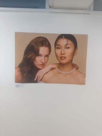 The photograph displays a close-up portrait of two young women against a plain, light brown backdrop. The woman on the left has long, wavy auburn hair and a fair complexion, with her gaze directed slightly away from the camera. Her left arm is draped over the shoulder of the other woman. The woman on the right has dark, straight hair pulled back from her face, with a more direct gaze towards the camera. She is wearing a double strand pearl necklace and a small stud earring. The image is presented as part of an exhibition, with a small white label affixed to the bottom left of the photograph. This label contains text in a small, dark font, with the first line appearing to read "Emily Richer" and the second line likely detailing the work's title or artist's specialization, such as "BA Makeup and Hair Design." The overall setting suggests an indoor exhibition space, possibly a gallery or a public venue, with a hint of a white ceiling and a dark horizontal band visible at the very top of the frame. Without further context regarding the exhibition itself or its location, it is impossible to definitively place this scene within Epsom and Ewell, United Kingdom.