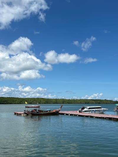 The image captures a scenic view of a body of water, likely a bay or strait, with a lush green treeline forming the background. A wooden pier extends into the water, accommodating two boats. The boat on the left is a traditional longtail boat, painted in brown and blue with an orange trim, and features a yellow flag and a Thai flag. It is docked at a floating platform labeled "ANDAMAN". To its right, a modern white speedboat with blue accents is moored to the pier. The speedboat has the text "9 C. CANOEING TOUR" visible on its side. The water is calm with gentle ripples, reflecting the bright blue sky dotted with fluffy white clouds. The overall atmosphere is serene and suggests a tropical location, consistent with Tambon Ko Panyi, Thailand, known for its floating villages and coastal scenery. No people are visible in the image.