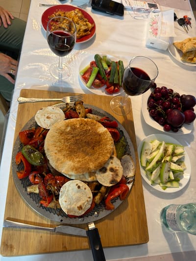 A bird's-eye view captures a table laden with food and drinks, likely set for a meal in Even Yehuda, Israel. The spread includes plates of cherries and plums, sliced zucchini seasoned with pepper, a red plate of baked pasta or casserole, and a serving dish of grilled vegetables with pita bread. Two glasses of red wine are present, one partially obscuring the pasta dish, and a bottle of water with Hebrew text sits nearby. A cutting board holds the grilled vegetables and pita. Other items on the table include a roll of paper towels, a set of keys, a remote control, reading glasses, and a smartphone. The setting appears to be indoors, with a bright, diffused light suggesting daytime. The overall impression is one of a leisurely gathering and a shared meal. The Hebrew text on the water bottle can be partially deciphered as containing information about deposits, suggesting a possible local consumer product.