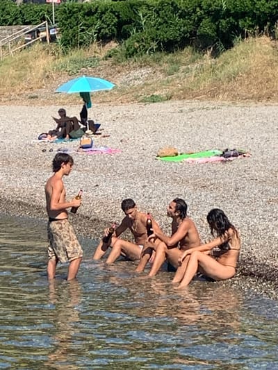 Four individuals are present on a pebbled beach in Pozzarello, Italy. In the foreground, partially submerged in clear water, a young man stands with a bottle in his hand. To his right, two men and a woman are seated on the shore, also with drinks. In the background, under a bright blue umbrella, two people are lying on towels. The environment is a sun-drenched beach with lush green vegetation lining the background hills.