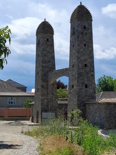 The image displays two identical stone towers, connected by an arched walkway. The towers have a tiered, pyramidal roof, topped with a pointed finial. Small rectangular openings are visible on the towers. The towers are constructed from large, dark gray stone blocks. The walkway between the towers is also made of stone and features a decorative, crenelated edge.
In the background, to the left of the towers, is a low-rise building with a grey tiled roof and a brick wall. Further to the left, a tree with green leaves is partially visible. To the right of the towers, another building with a wooden roof structure is present. The foreground is occupied by a dirt path paved with cobblestones, and an area of tall, green vegetation, some of which is dry and brown. The sky is cloudy with patches of blue. The scene appears to be an outdoor architectural site, possibly a historical monument or a newly constructed structure designed to resemble historical architecture. The location is identified as the city of Urus-Martan, Russia.