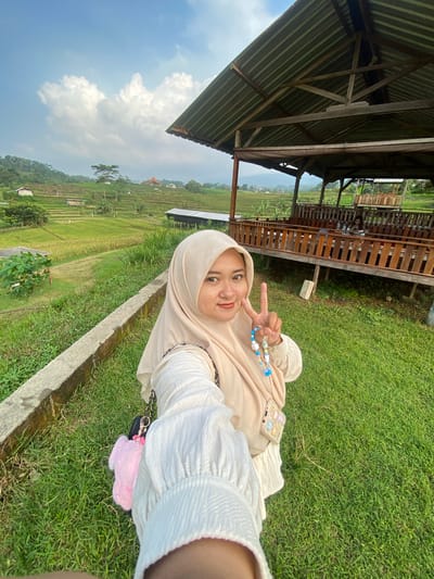 A young woman in a beige hijab is taking a selfie in the city of Penanggungan, Indonesia. She is making a peace sign with her fingers. She is wearing a light-colored top. A beaded bracelet is visible on her wrist. She has a phone with a decorative case placed on her lap. She is sitting on a grassy area. Behind her is a wooden structure with a roof, and beyond that, a terraced rice field landscape is visible under a partly cloudy sky. A concrete structure or border runs along the edge of the grass where she is sitting. A pink fluffy object attached to a black purse chain is visible in the lower right.