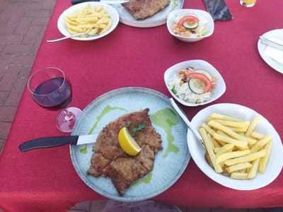 A table is set for a meal on a red tablecloth. In the foreground, a large schnitzel sits on a light blue plate, garnished with a lemon wedge and parsley, and a knife is placed on the plate. To the right, a bowl of french fries is visible, with a spoon inside. A wine glass filled with red wine is to the left of the schnitzel. In the background, there are two small bowls of salad with cucumber and tomato slices, and another plate with a schnitzel and fries. A mobile phone is also on the table. The setting appears to be an outdoor restaurant in Monheim am Rhein, Germany, with a cobblestone patio visible in the upper left corner of the image.