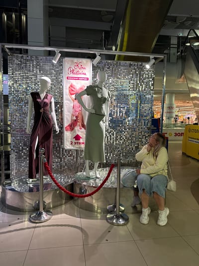 In Ermakovo, Russia, a woman is seated in front of a retail display featuring two mannequins dressed in women's clothing. The display is against a shimmering, sequined backdrop. The mannequin on the left wears a dark, floor-length dress with a high slit and a gold accent. The mannequin on the right wears a light green dress with a collar. A pink banner between the mannequins reads "Kira Plastinina" and "ЖЕНСКАЯ ОДЕЖДА" (Women's Clothing) along with an arrow indicating "2 этаж" (2nd floor). A red rope barrier is in place in front of the display. The woman, wearing a light yellow long-sleeved shirt, denim shorts, and white sneakers, is looking upwards at the display. She has a white purse over her shoulder and is holding a plastic bag. The surrounding environment appears to be a shopping mall, with escalators and other retail signage visible in the background.