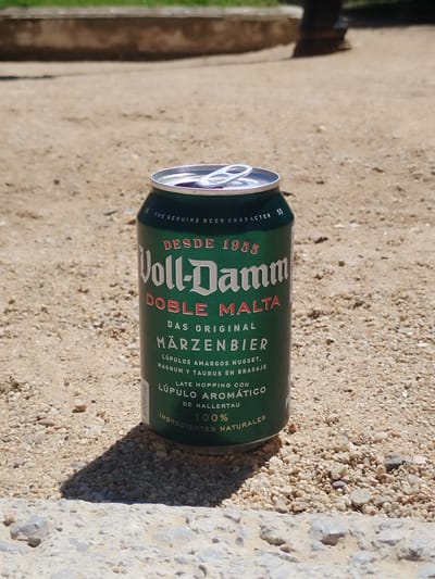 A can of Voll-Damm Doble Malta beer sits on a sandy, pebbly surface, likely outdoors. The can is green with white and red lettering and has been opened. The background is blurred, showing hints of greenery and possibly a building or structure. The can's shadow is cast on the ground. The location is unspecified; Rubí, Spain, is not visibly confirmed within the image.