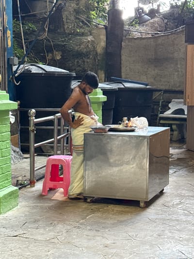 This image captures a man engaged in a religious or ceremonial activity, likely at a shrine or temple located within the Selayang Municipal Council area in Malaysia. The scene is set outdoors or in a semi-open space, illuminated by natural daylight.

The central subject is a man of South Asian descent, dressed in traditional attire. He wears a light-colored dhoti or veshti wrapped around his waist, leaving his chest bare, and has multiple strands of mala beads around his neck. He has dark hair and a beard, and his posture suggests focus or reverence as he stands behind a rectangular, silver-colored metal counter or cabinet on wheels. His hands are positioned in front of him.

On the surface of the metal counter, several items are placed, including a small metal lamp (diya) with a visible flame and what appears to be white cloth or offerings. A red plastic stool is visible to the man's right, next to the counter and metal railings. The ground is a textured concrete surface, showing signs of wear and cracking in places.

The background features natural elements, including large rock formations partially covered with green foliage and vines, suggesting a integration with the natural environment. A black speaker is visible in the mid-ground, and various electrical cables are strung across the scene. Other elements include green-painted concrete structures and dark industrial-looking objects. No discernible text is visible in the image. The overall impression is one of a spiritual or ritualistic event unfolding in a humble yet dedicated setting.