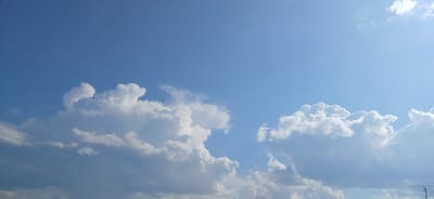 The image depicts a clear blue sky with scattered cumulus clouds. The clouds are white and fluffy, with some appearing darker and more dense, suggesting varying atmospheric conditions. The lighting indicates it is daytime, with sunlight filtering through the atmosphere. The horizon is not visible, as the image is focused on the sky. There are no people, animals, or man-made structures clearly identifiable in the frame, with the exception of a faint metallic structure on the far right, possibly an antenna. The sky is a vibrant blue, transitioning to a lighter shade towards the upper right corner where the sun may be located. The image is a typical representation of a sky on a clear day.
