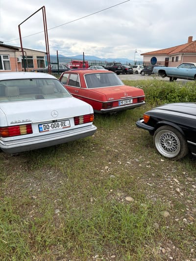 In Natakhtari, Georgia, on a cloudy day, several vintage cars are parked on a gravel and grass lot. In the foreground, a white Mercedes-Benz 420 SEL with a Georgian license plate "DO-008-RE" is visible. To its right, a red Mercedes-Benz 230 with the Georgian license plate "MR-230-MR" is parked. A black Mercedes-Benz is partially visible to the right of the red car. In the background, a red fire truck is parked near a building with an orange and beige facade and a chain-link fence. Further in the background, other vehicles, including SUVs and sedans, are parked, and a light blue convertible is seen. Mountains are visible in the distance. The scene appears to be an outdoor parking area, possibly near a commercial establishment or a transportation hub.