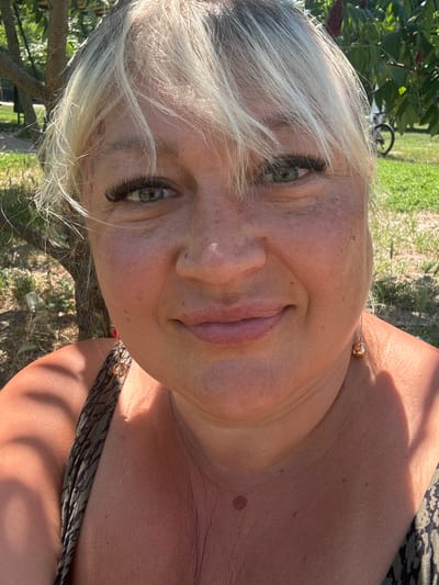 This is a close-up, vertically oriented, outdoor photograph of a woman. She has blonde hair, green eyes, and fair skin with freckles. She is wearing a dark, patterned garment, possibly a dress or swimsuit. A delicate gold necklace with a sphere pendant is visible around her neck. The woman is looking directly at the camera with a relaxed, pleasant expression.
The background suggests a natural setting, with out-of-focus green foliage and grass, hinting at a park or garden. The lighting indicates it is daytime, likely sunny, with shadows visible on her skin, suggesting the sun is at an angle. The overall mood is casual and warm. The image was taken in Zmiiv, Ukraine, though no specific landmarks are identifiable. There is no visible text in the image.