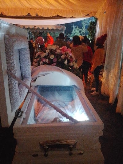 The image depicts a funeral scene occurring under a draped canopy in Marenyo-Shianda ward, Kenya, during evening or night hours. In the foreground, a white, textured coffin with an open lid is positioned. The coffin features ornate metal handles and a transparent viewing panel on its top surface. Inside, a body covered in light-colored fabric is visible, illuminated by a bright light source within the coffin's lower right interior. A braided, light-colored rope-like object extends diagonally across the interior of the open lid and over the viewing panel. A large floral arrangement, predominantly pink, white, and purple, rests on the partially closed section of the coffin. In the mid-ground, a gathering of approximately 7-10 individuals, whose features are indistinct, are present. One individual wears a bright red top, while another on the far right wears a yellow-orange top and a matching beanie hat. In the background, an additional white tent structure and trees are visible against a dark sky.