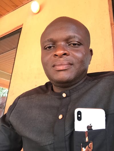 A dark-skinned male individual, appearing middle-aged and bald, is depicted in a close-up, head-and-torso shot, facing forward with a neutral expression. He is wearing a dark, possibly black, long-sleeved traditional-style shirt featuring a band collar and two visible copper-toned buttons on the placket. The shirt has textured fabric panels across the shoulders and upper chest. A white smartphone, identified as an iPhone with dual rear cameras and an Apple logo, is partially inserted into the shirt's left breast pocket. A small section of a brown and black patterned fabric is visible beneath the phone within the pocket. The background consists of a plain, light orange-colored wall. An illuminated, spherical light fixture is mounted on the wall to the upper left of the individual's head, and a partial dark-framed window or door with light-colored paneling is visible below the light. The scene is located in Adum Afo, Nigeria.