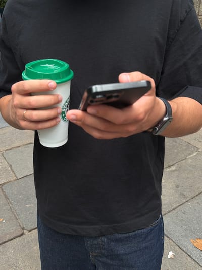 In Santiago De Compostela, Spain, a person stands on a cobblestone pathway, holding a Starbucks coffee cup in their left hand and a smartphone in their right. They are wearing a black t-shirt and blue jeans. The coffee cup has a green lid and the Starbucks logo is visible. The person is looking at the smartphone. The background shows a cobblestone street and part of a building.