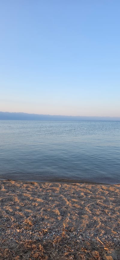 A serene, wide-angle vertical shot captures the tranquil scene at the shores of Issyk-Kul lake, near the village of Boz Beshik, Kyrgyzstan. The image is divided into three horizontal bands: a clear, pale blue sky occupies the top third, transitioning to a soft, hazy horizon line of distant mountains in muted blues and purples. The middle section is dominated by the calm, glassy surface of the lake, reflecting the sky and distant landscape with subtle ripples near the shore. The foreground consists of a pebbly beach, illuminated by warm, low-angled sunlight casting dappled shadows and highlighting the texture of the stones and scattered dry vegetation. There are no people or explicit actions depicted, emphasizing the natural, undisturbed beauty of the environment. The overall atmosphere is one of peacefulness and stillness, typical of early morning or late afternoon light.