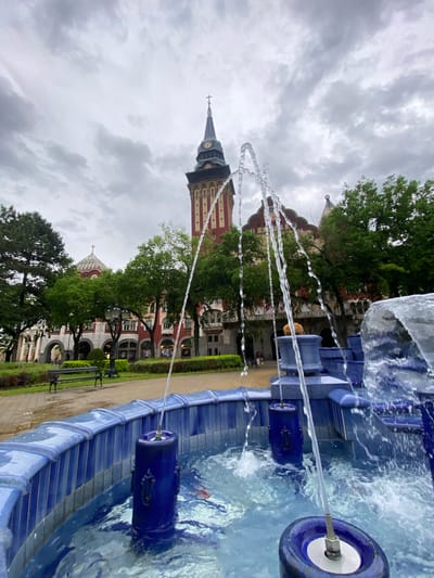 The image captures a water fountain in Subotica, Serbia, with the City Hall tower visible in the background. The fountain is blue and features decorative cylindrical elements from which water spouts. The City Hall is a tall, ornate structure with a clock tower and a spire topped with a cross. It's partially obscured by trees. A path with benches runs alongside a grassy area. The sky is overcast with gray clouds. The overall perspective is slightly tilted.