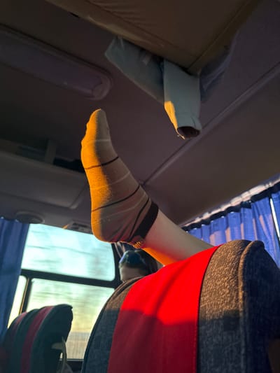The image appears to be taken inside a vehicle, possibly a bus or train. A person's leg and foot, wearing a striped sock, is extended towards the aisle. The person seems to be sitting by the window. The window shows a blurred view of the outside. The interior has a luggage rack with a pillow, and curtains that appear blue. The image does not contain any information confirming the location is Simakovo, Russia.