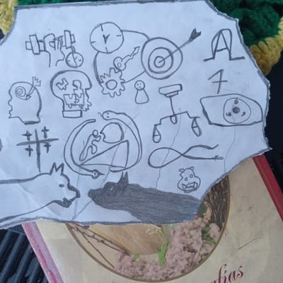The image displays a torn piece of paper with various hand-drawn sketches. These sketches include a profile of a head with gears and a spiral inside, a skull, a person lifting weights, a clock connected to a target with a key and a gear, a stick figure, a set of scales, a snake coiled around a person, an infinity symbol, a stylized eye with three circles, a tic-tac-toe grid, a dog-like creature, and a quadrupedal animal, possibly a wolf or dog, depicted in dark gray shading. The paper is placed on top of a book with a floral pattern visible on its cover. The background suggests an outdoor setting with a knitted object in green and yellow colors. The overall impression is of a collection of symbolic or conceptual drawings. There are no discernible elements that indicate a specific location in Duaca, Venezuela.