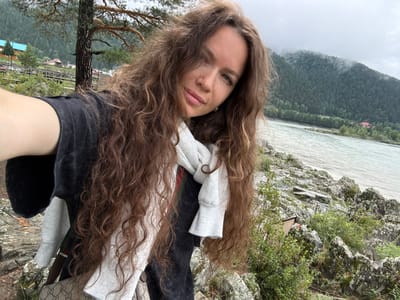 A young woman with long, curly brown hair is taking a selfie outdoors. She is wearing a black t-shirt and a white scarf. Her left shoulder and arm are visible in the foreground. The background shows a river, a rocky shore, a forest-covered mountain range, and a small village with blue and red-roofed buildings. The sky is overcast. The location appears to be Elekmonar, Russia.