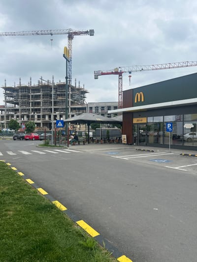 The image shows a McDonald's restaurant next to a construction site. The restaurant has a McCafé section and a handicapped parking space. Two cranes are visible in the background next to the construction site. Several parked cars are in the foreground and background near a marked pedestrian crossing. The location is likely in Giurgiu, Romania, based on the store signage. The sky is overcast.